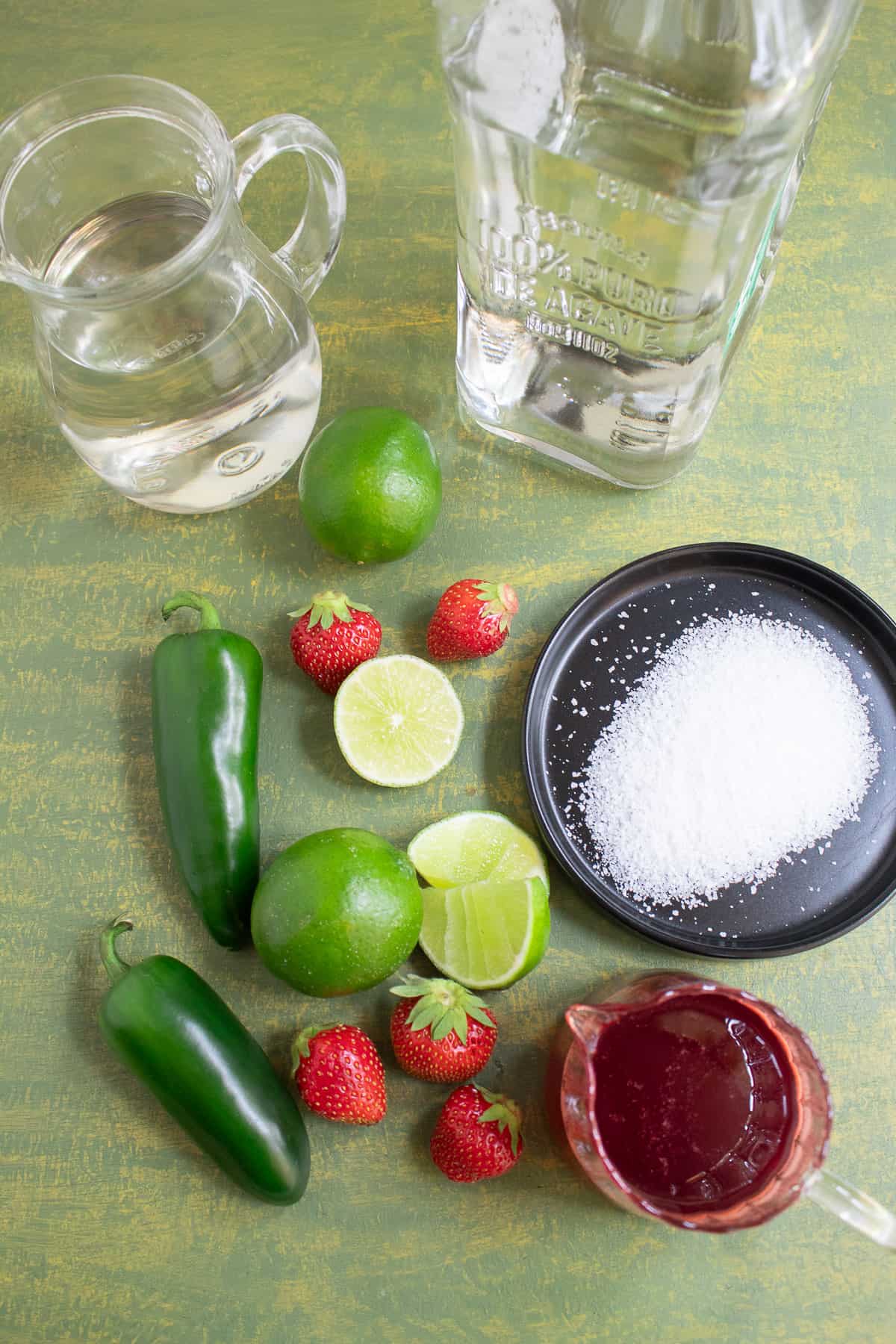 Ingredients for the strawberry jalapeno margaritas arranged on a green surface.