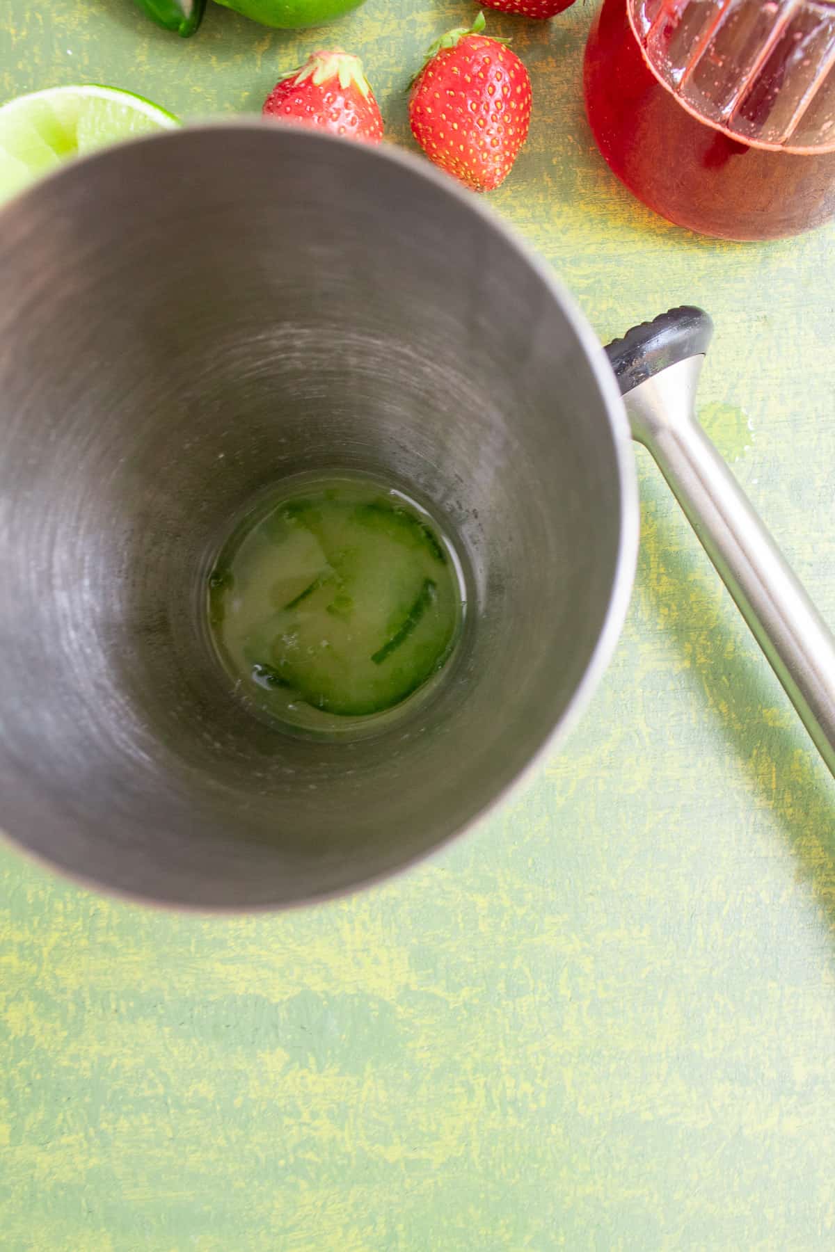 A top-down view into a cocktail shaker with jalapeno slices and lime muddled together. On the green surface next to the shaker sits a cocktail muddler, fresh strawberries, and lime wedges.