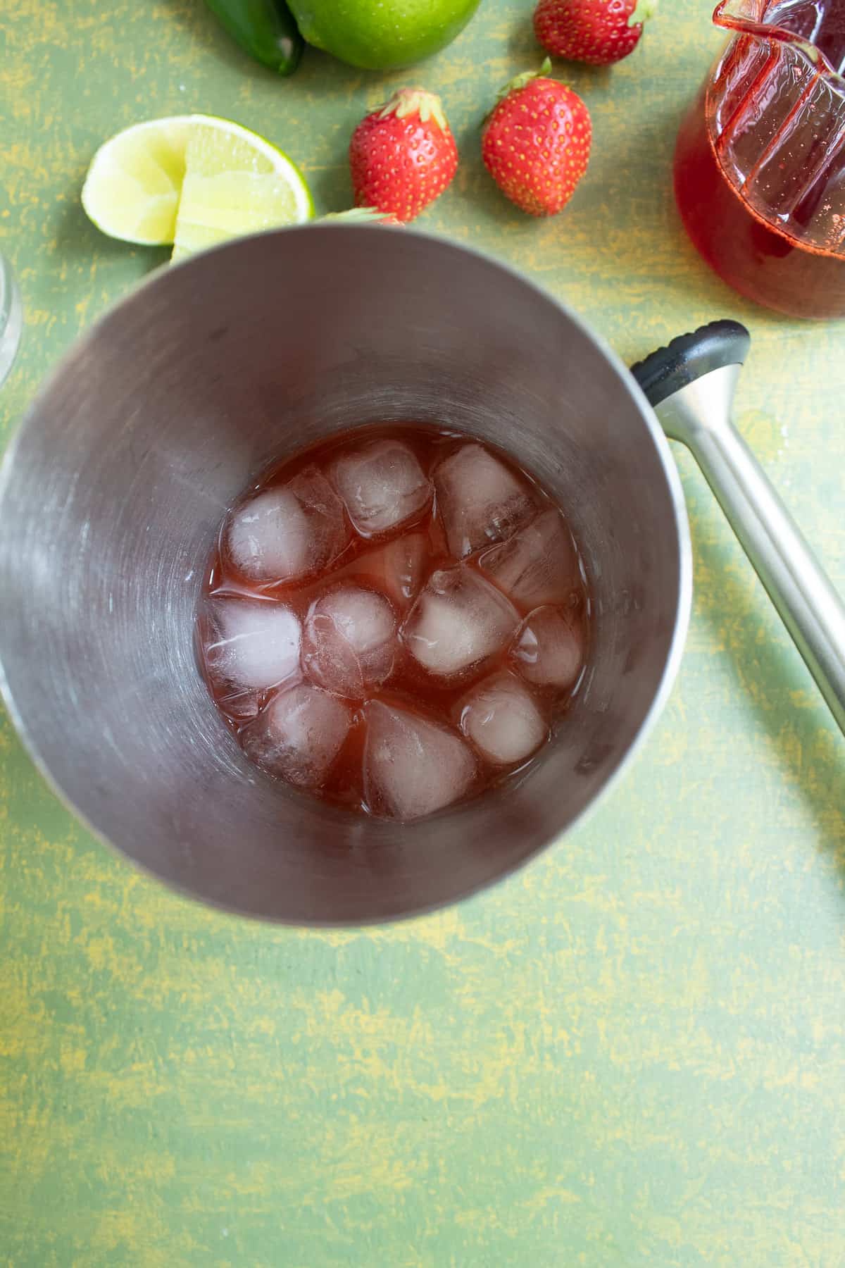 A top-down view into a cocktail shaker with small ice cubes and a red liquid. On the green surface next to the shaker sits a cocktail muddler, fresh strawberries, and lime wedges.