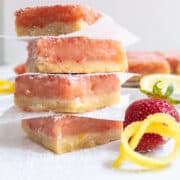 Pink dessert bars are stacked on a white plate with lemon peel and strawberry slices as garnish.