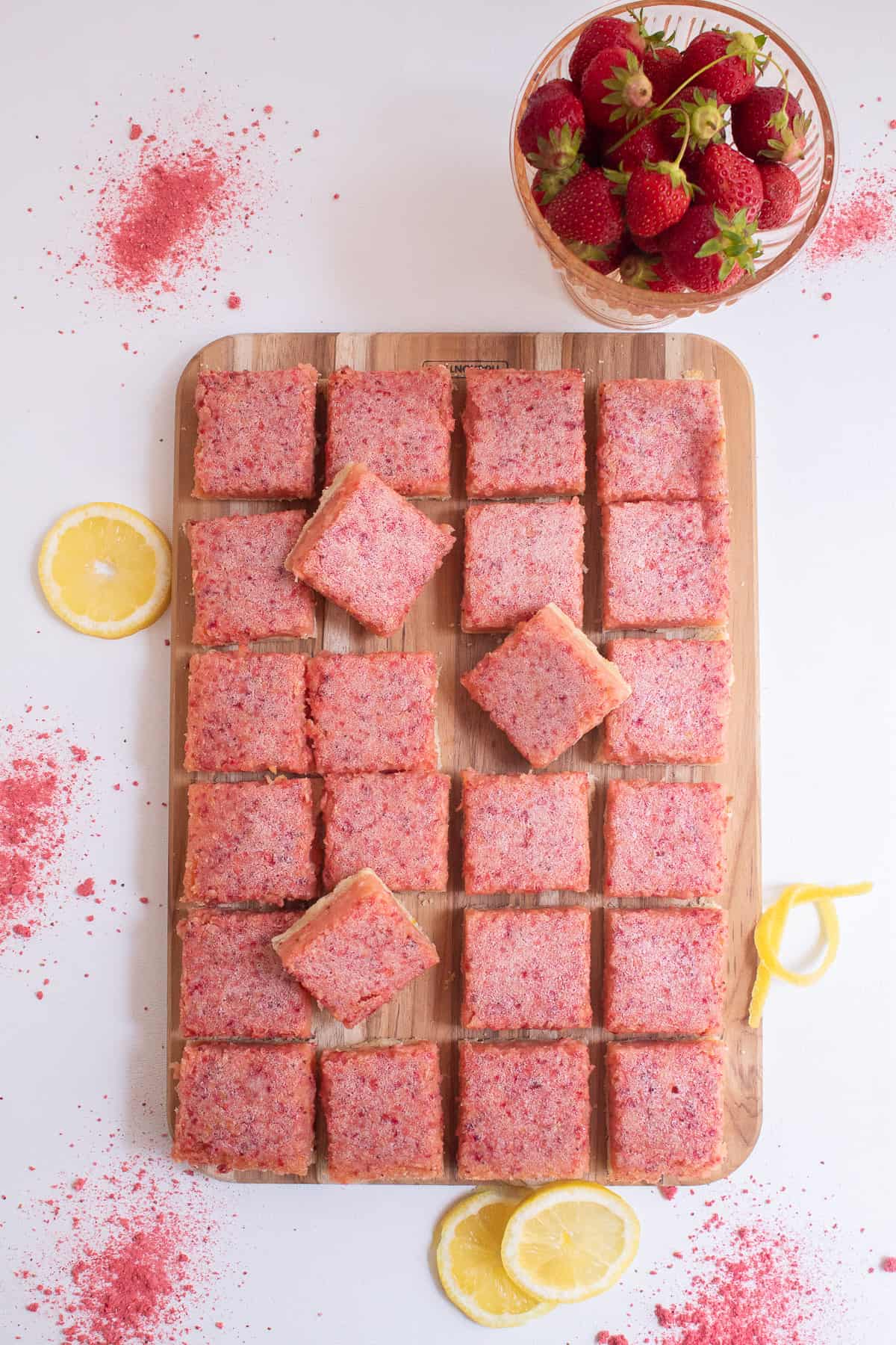 The strawberry lemon bars are on a wooden cutting board ans are surrounded by fresh strawberries, lemon slices, and strawberry powder.