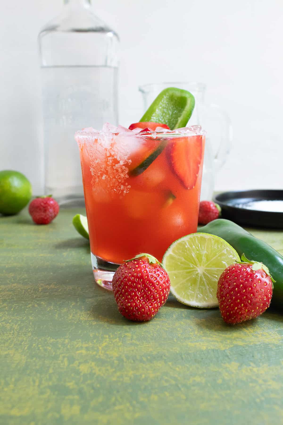 A rocks glass with a salted rim holds a red liquid and is garnished with a sliced strawberry and a slice of jalapeno. In fornt of the glass are two fresh strawberries, a sliced lime, and a fresh jalapeno.