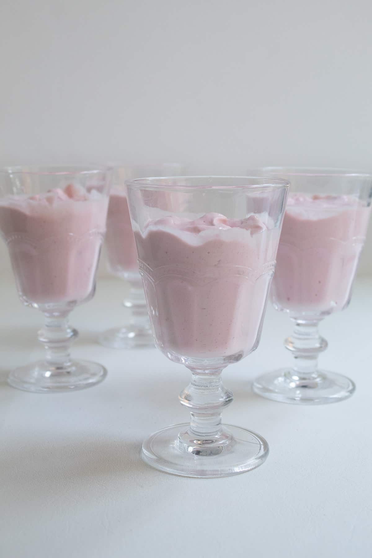 The strawberry mousse is divided among four footed serving glasses sitting on a white surface.