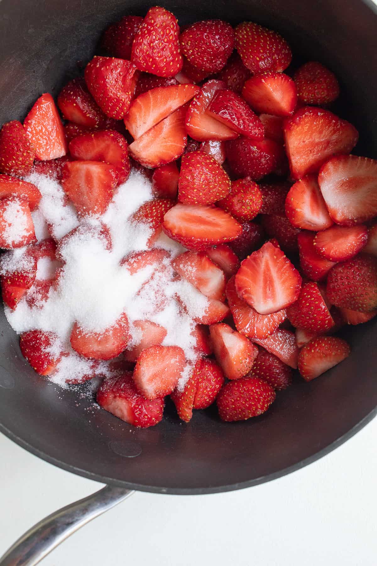 A black saucepan contains sliced strawberries and sugar.