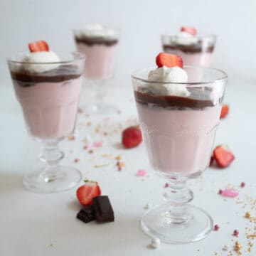 Footed serving glasses are filled with a layer of pink mousse, a layer of dark chocolate, and a garnish of whipped cream and a sliced strawberry.