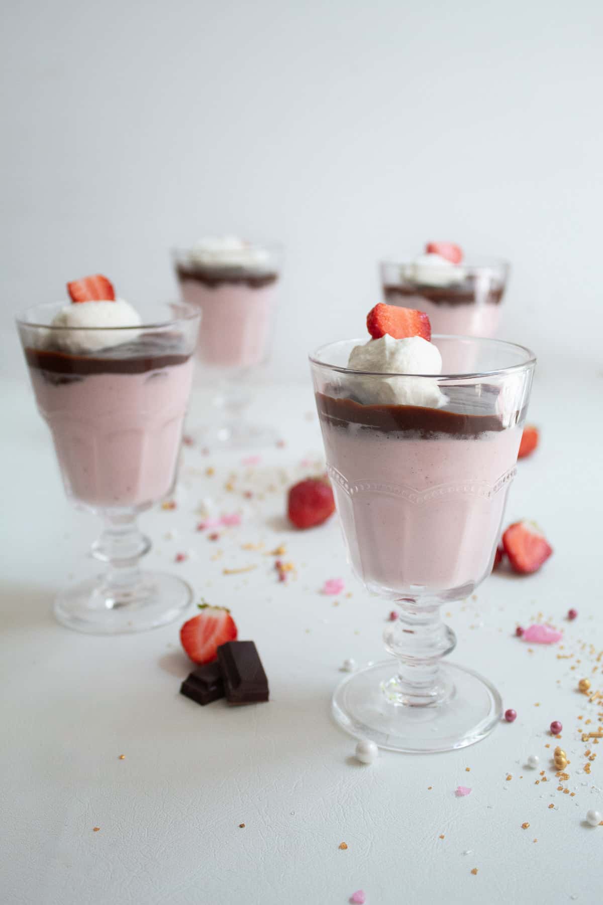 Footed serving glasses are filled with a layer of pink mousse, a layer of dark chocolate, and a garnish of whipped cream and a sliced strawberry.