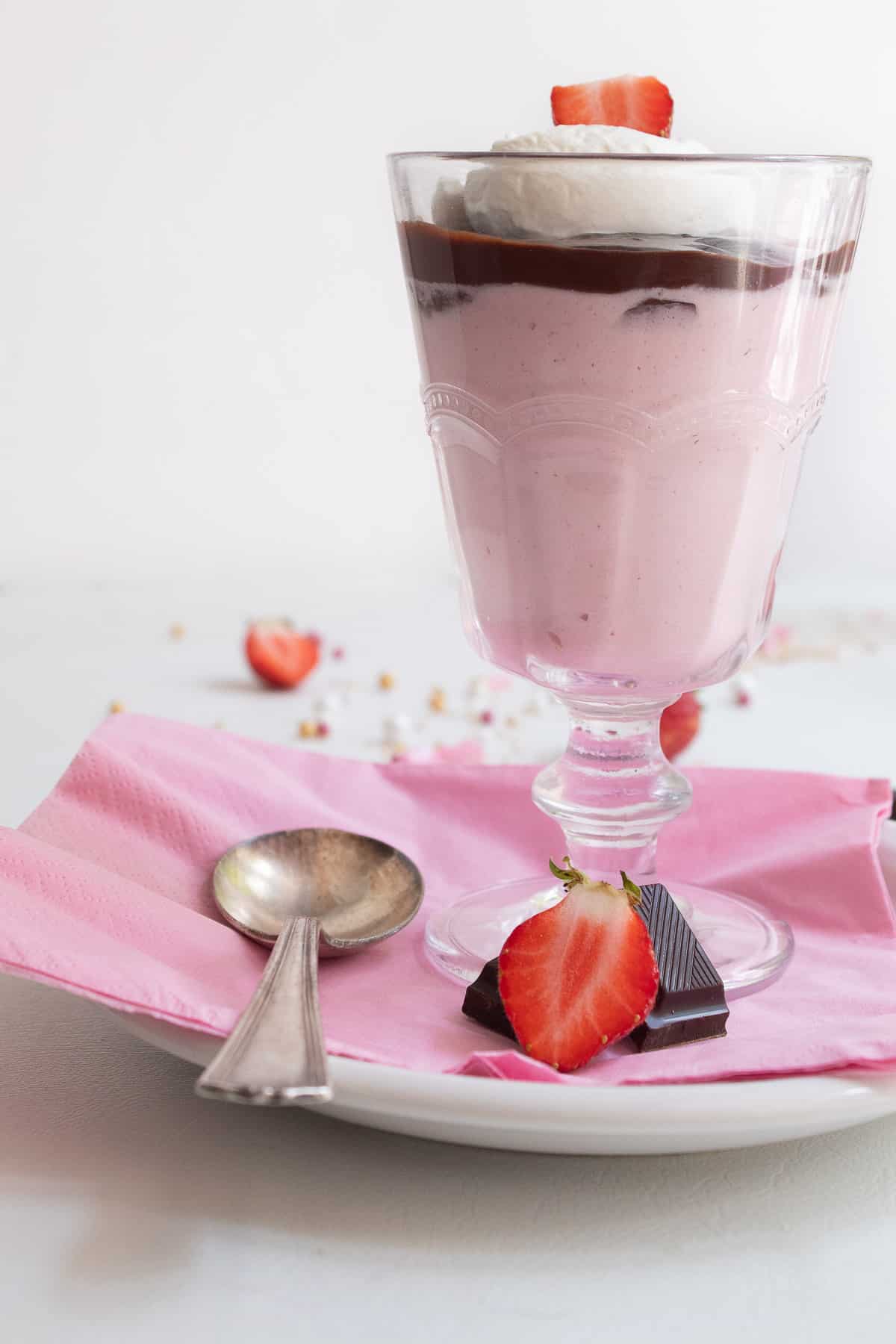 A footed serving glass is filled with a layer of pink mousse, a layer of dark chocolate, and a garnish of whipped cream and a sliced strawberry sits on a napkin-lined plate. A sliced strawberry and pieces of chocolate are in the foreground.