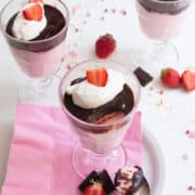 Three footed serving glasses are filled with a layer of pink mousse, a layer of dark chocolate, and a garnish of whipped cream and a sliced strawberry. A spoonful of the mousse sits on the serving plate in the foreground.