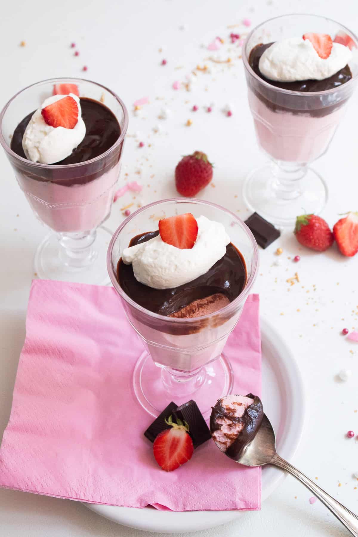 Three footed serving glasses are filled with a layer of pink mousse, a layer of dark chocolate, and a garnish of whipped cream and a sliced strawberry. A spoonful of the mousse sits on the serving plate in the foreground.