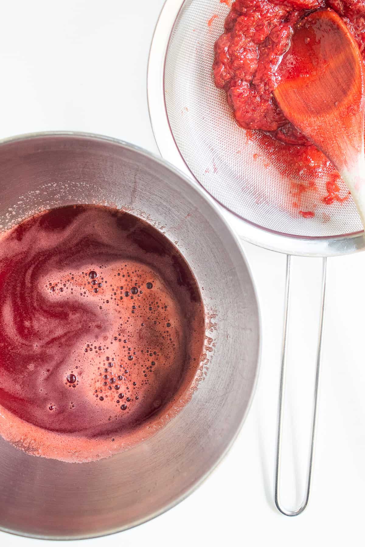 The strained strawberry mixture sits in a stainless steel bowl next to a strainer full of the strawberry solids.