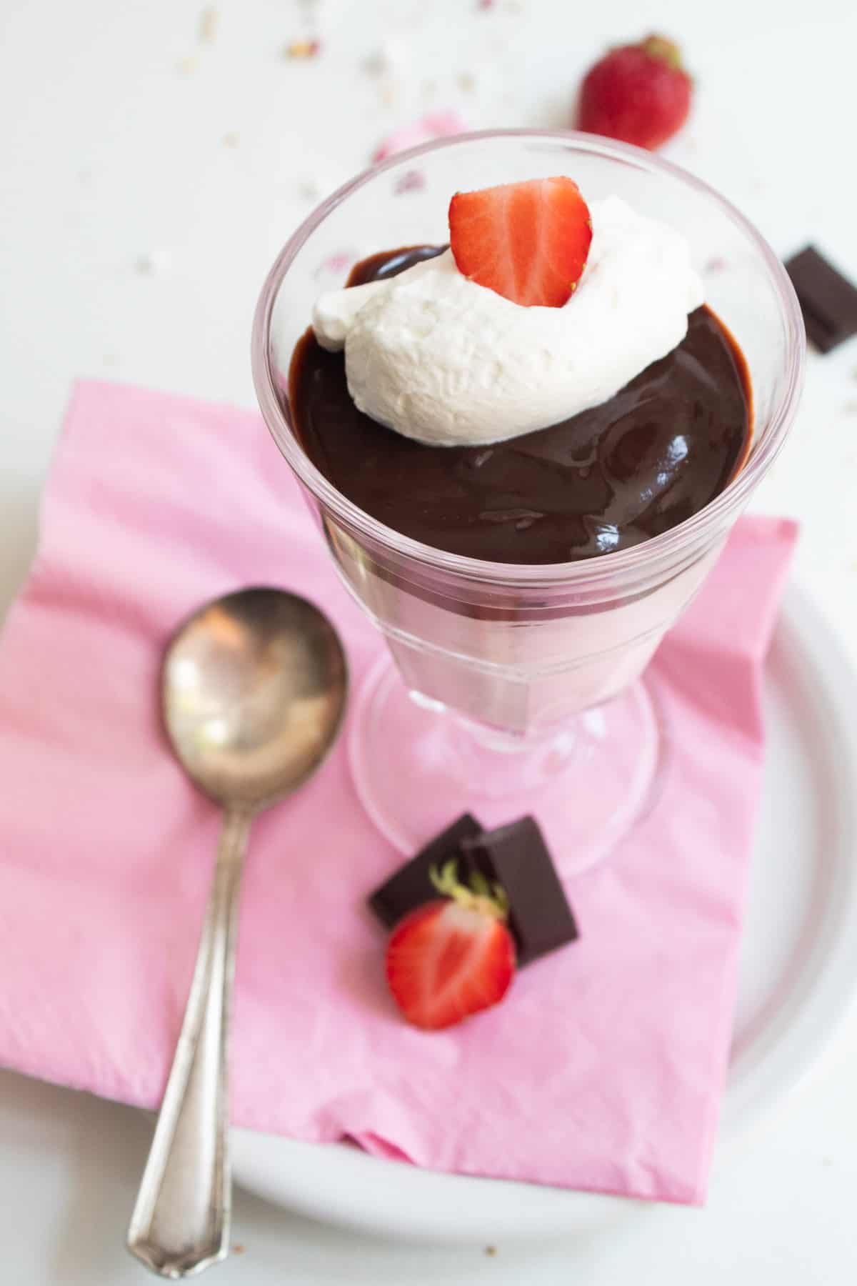 A footed serving glass is filled with a layer of pink mousse, a layer of dark chocolate, and a garnish of whipped cream and a sliced strawberry and sits on a plate lined with a pink napkin.