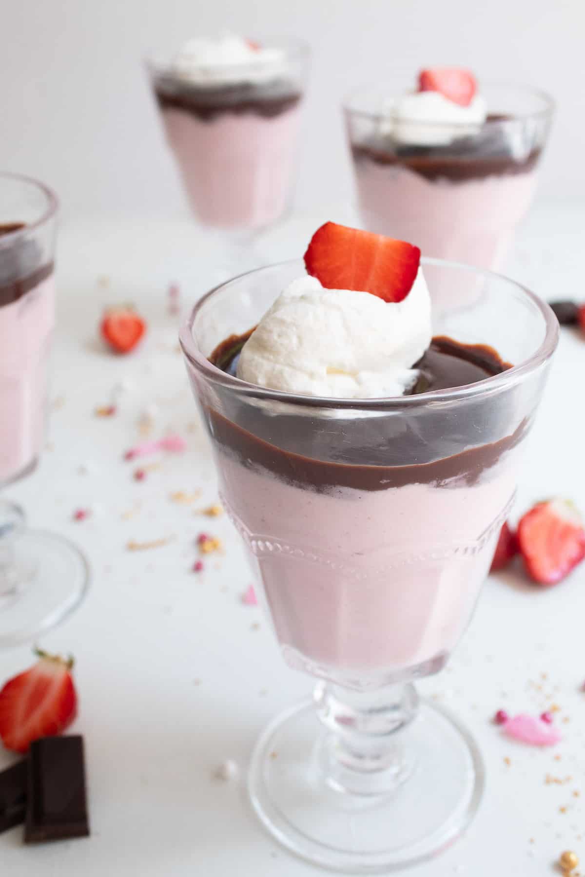 A footed serving glass is filled with a layer of pink mousse, a layer of dark chocolate, and a garnish of whipped cream and a sliced strawberry.