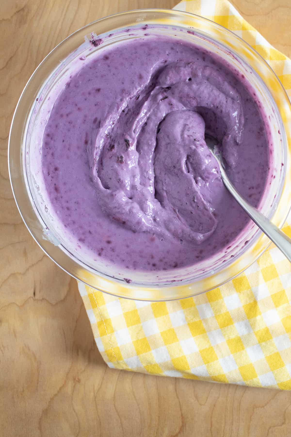 The chilled ice cream mixture is a rich purple color and sits on a light wooden surface in a glass bowl on top of a yellow checked napkin.