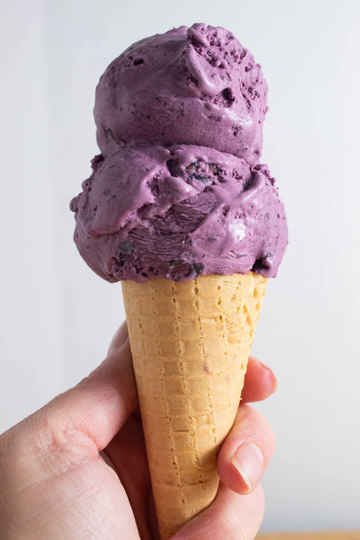 Two scoops of purple ice cream on a brown sugar cone being held by someone's left hand.