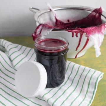 A jar of the blueberry syrup sits on a green and white striped towel next to a bowl used to strain the syrup.