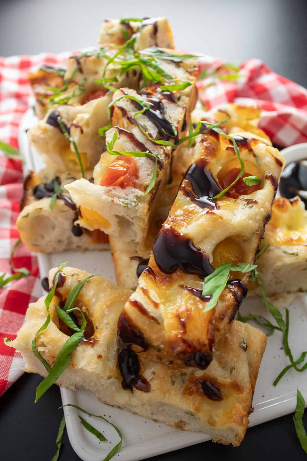 Slices of the focaccia stacked on a white tray and drizzled with balsamic glaze and shredded basil.