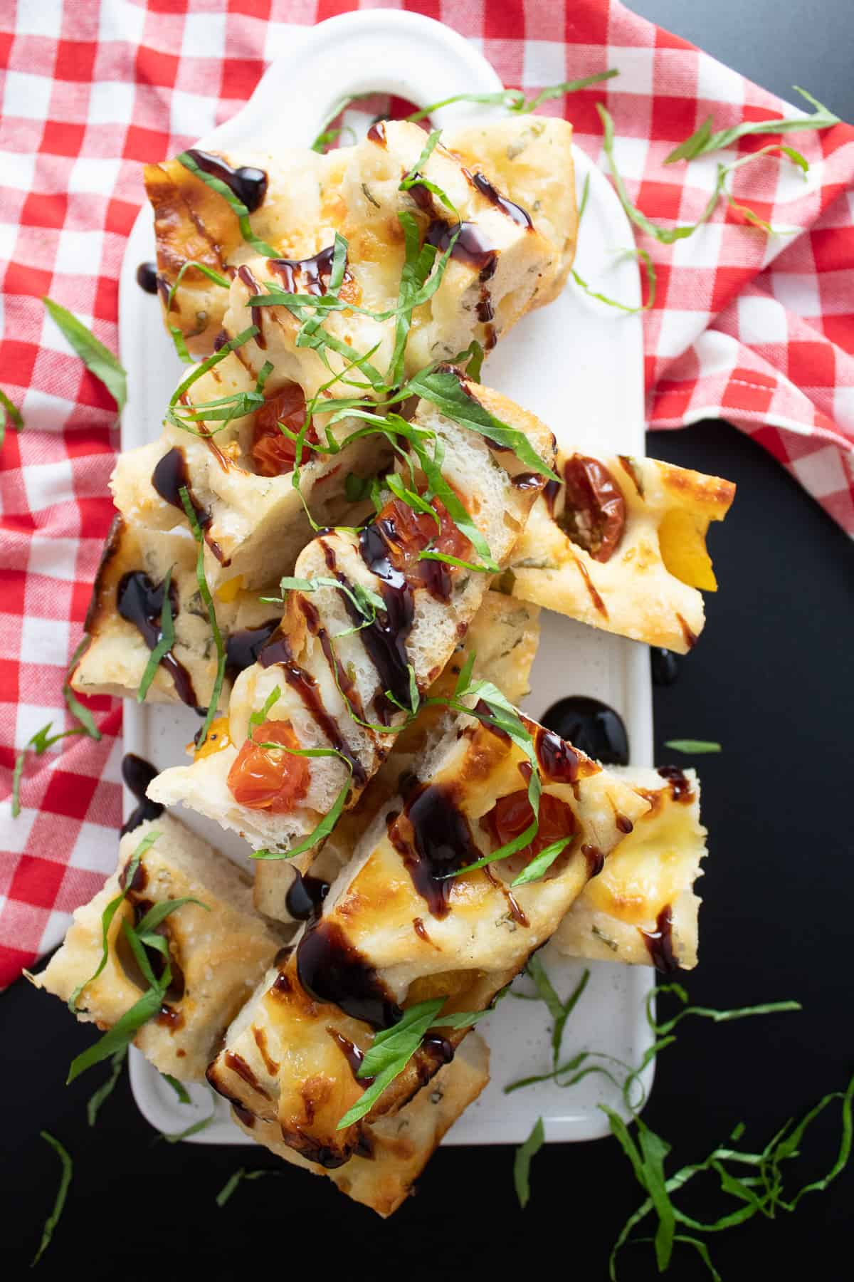 Slices of the focaccia are stacked on a white tray and drizzled with balsamic glaze and shredded basil.