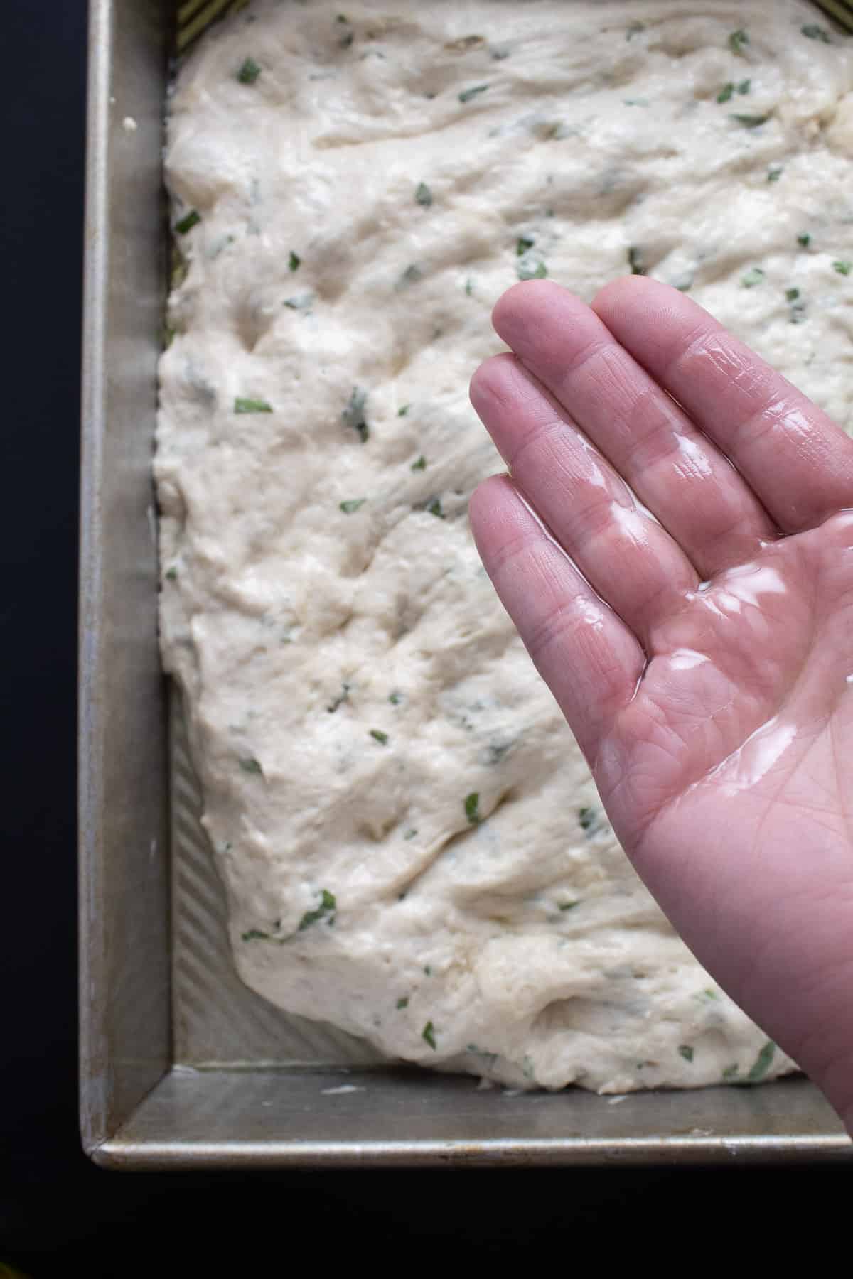 An oiled hand ready to flip the dough which rests in an oiled baking dish.