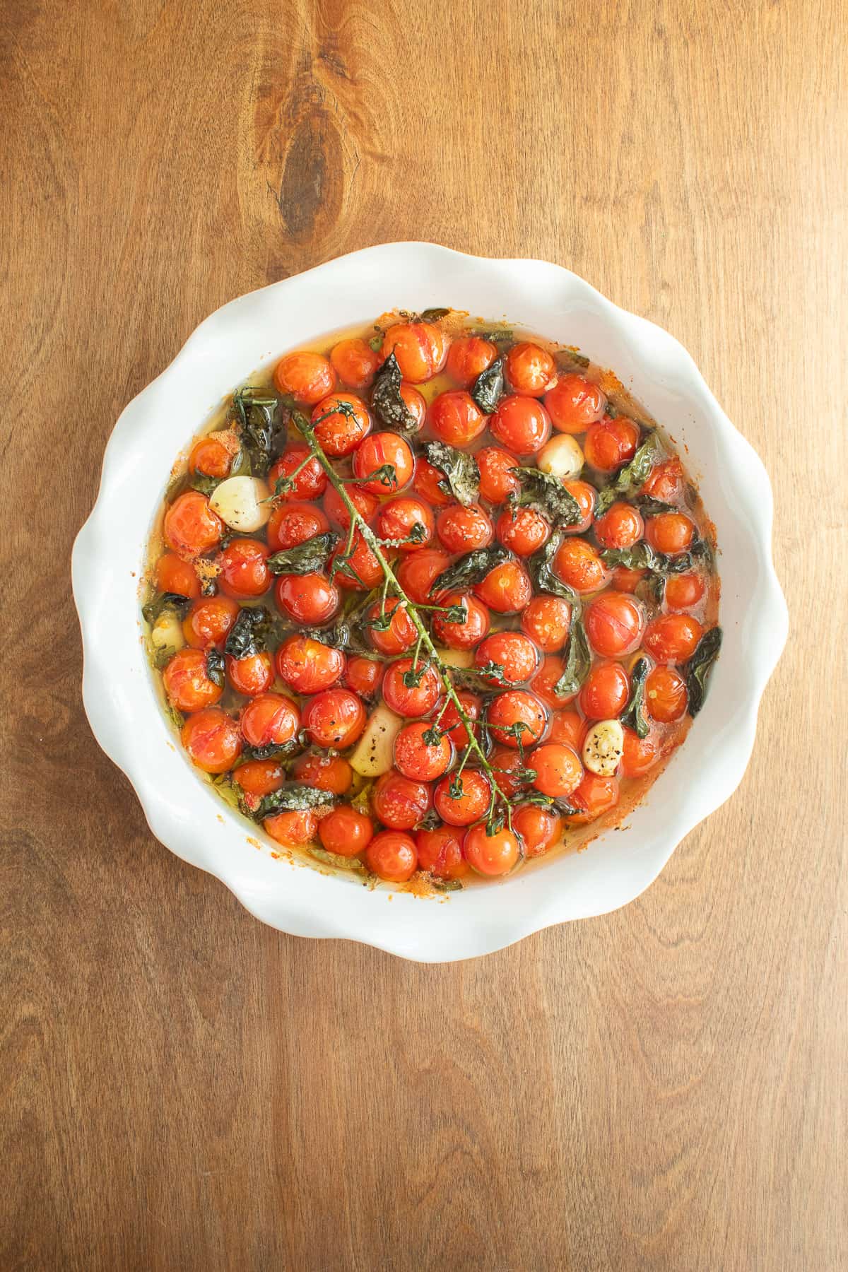 The roasted tomatoes, garlic, herbs, and oil in a deep white pie dish.