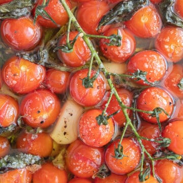 Burst cherry tomatoes on the vine sit in oil with pepper, garlic cloves, herbs, and salt.