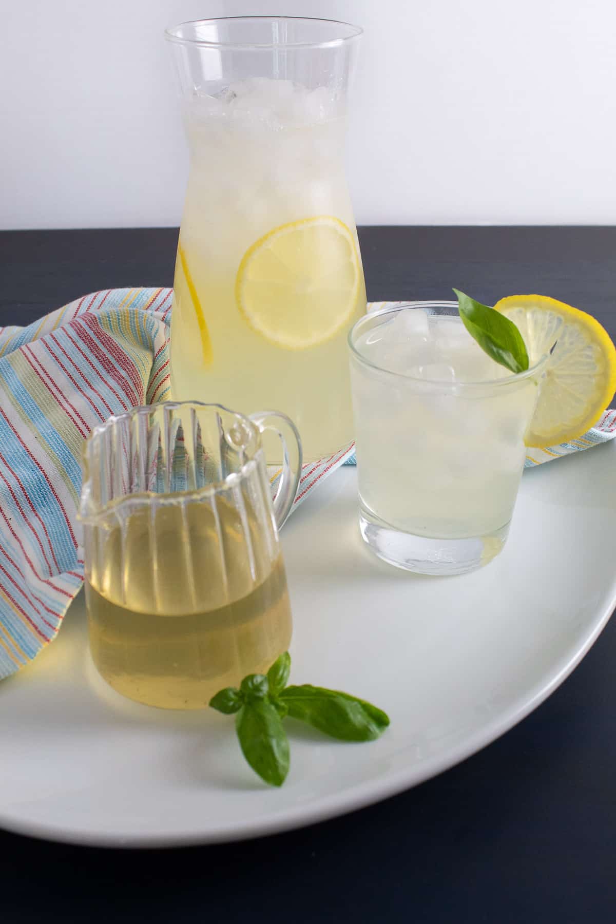 A pitcher of lemonade sits on a white tray next to a glass garnished with a basil leaf and a lemon slice and a small pitcher of basil syrup.