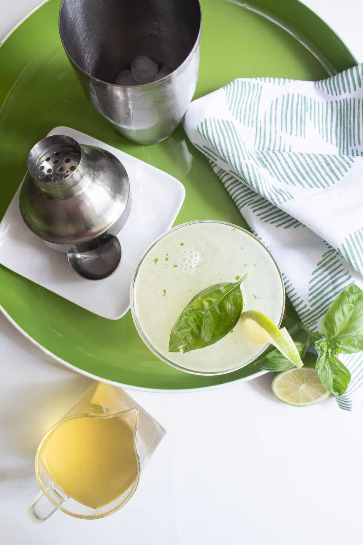 A pale green cocktail in a coupe glass sits on a green tray near a cocktail shaker, basil leaves, lime slices, and a pitcher of a pale yellow syrup.