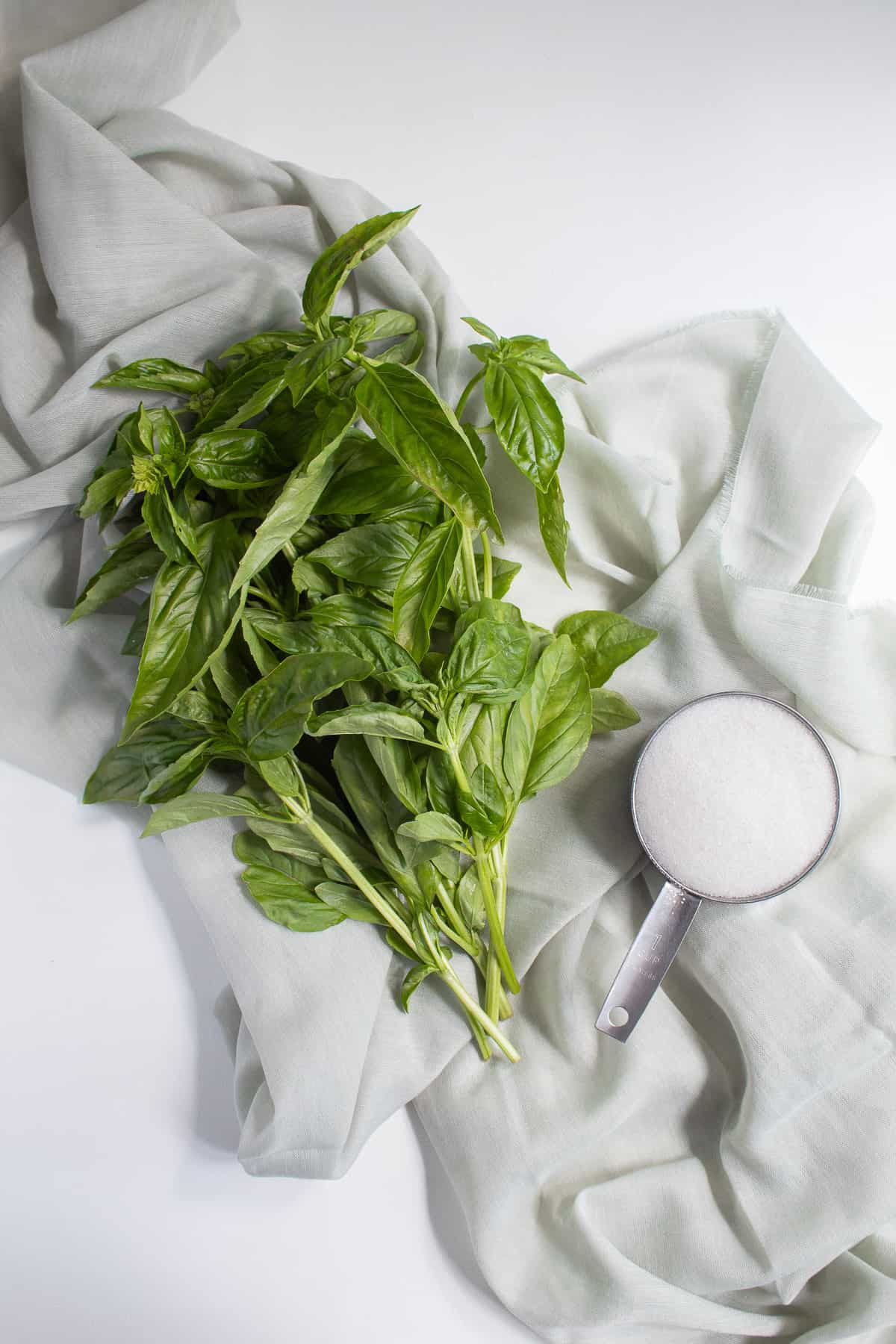 A bunch of basil leave sand a cup of sugar are displayed over a pale green fabric background.