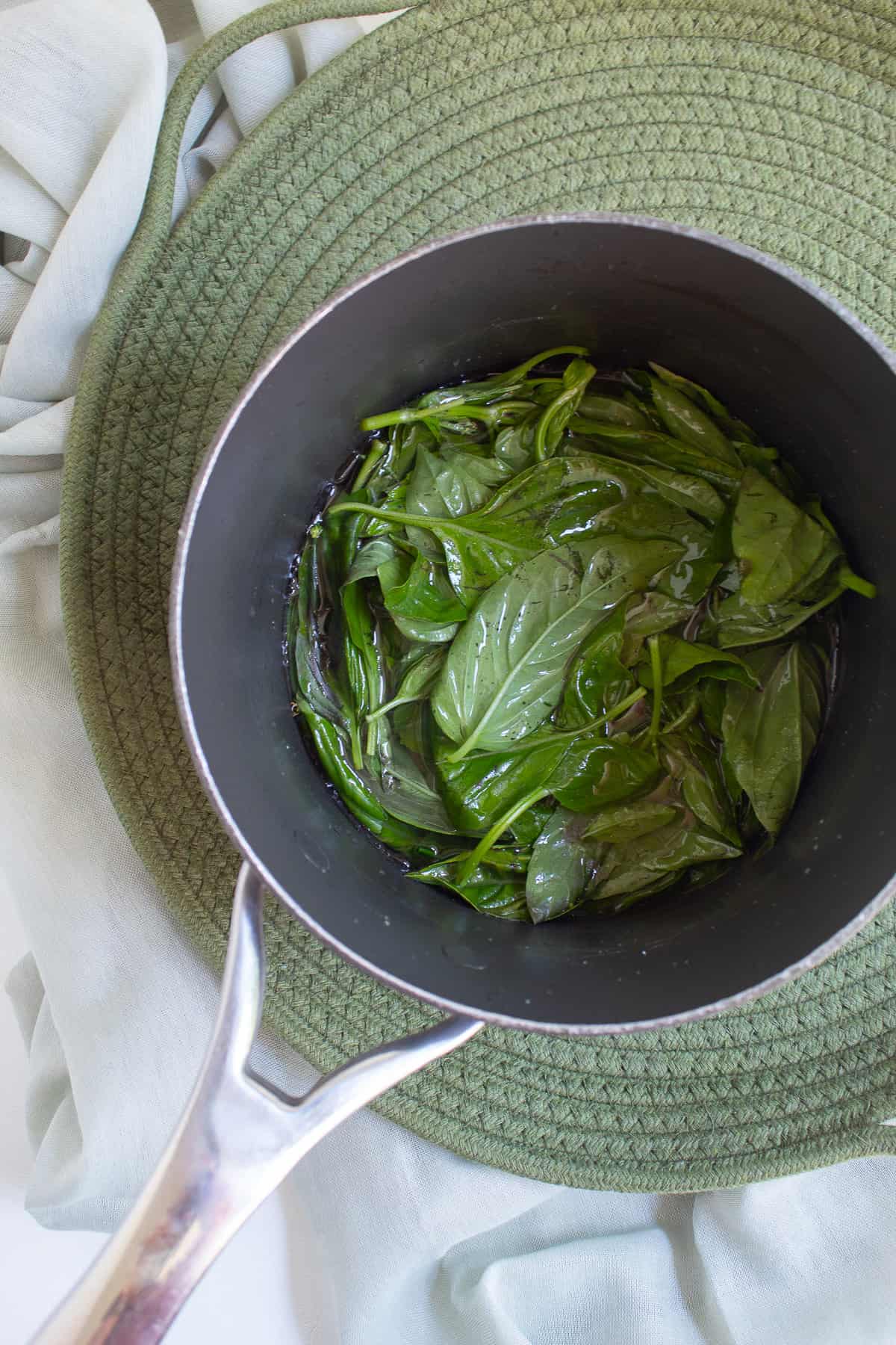 Basil leaves are submerged in a liquid in the bottom of a dark saucepan set on a green woven tray.