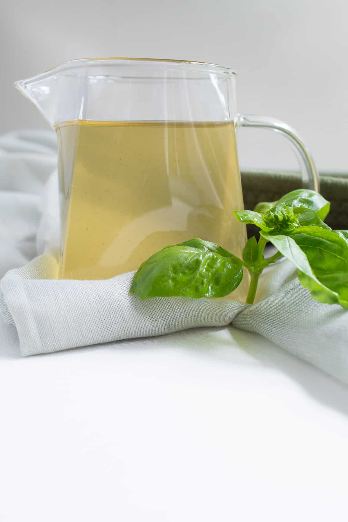A pale yellow syrup in a clear glass pitcher sits on green fabric next to basil leaves.