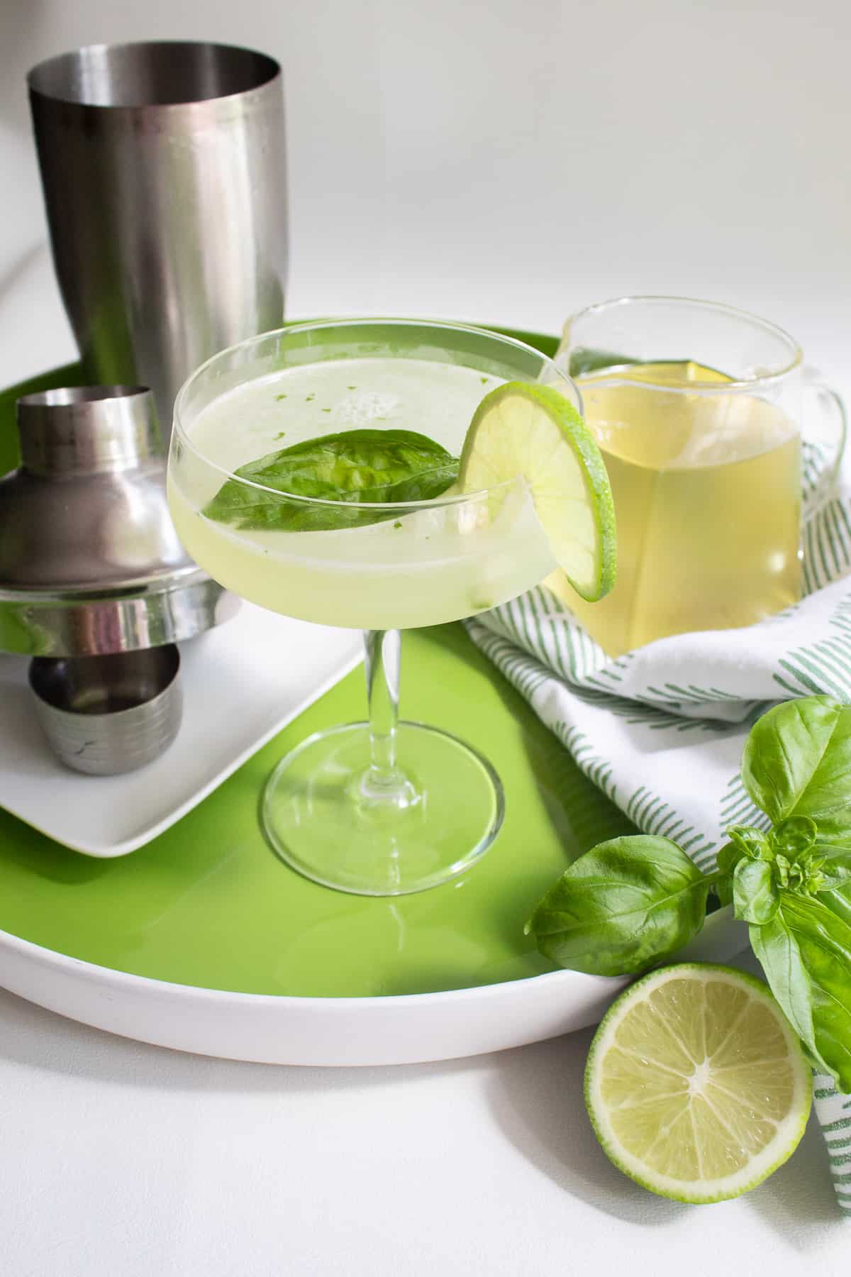 A pale green cocktail in a coupe glass sits on a green tray near a cocktail shaker, basil leaves, lime slices, and a pitcher of a pale yellow syrup.
