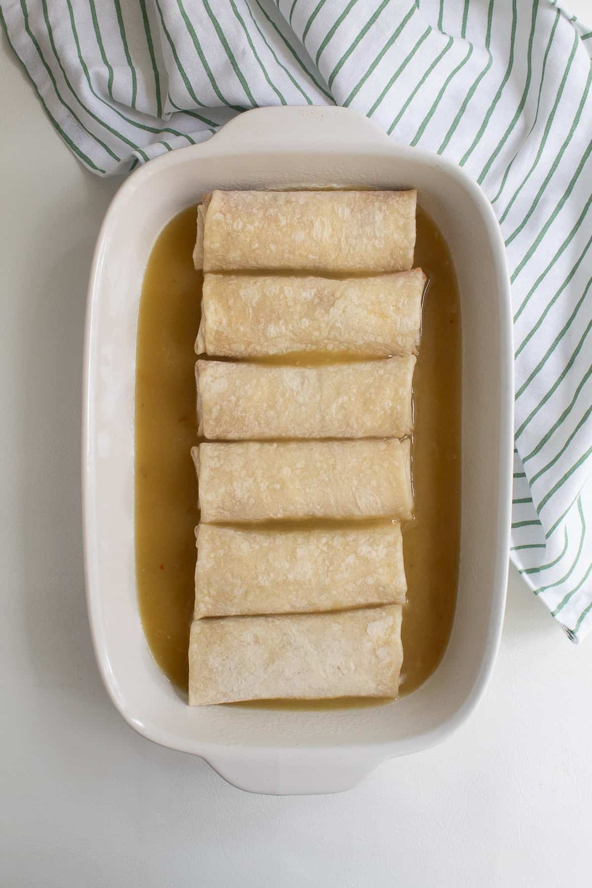 Six burritos are placed on top of a layer of green enchilada sauce in a casserole dish.