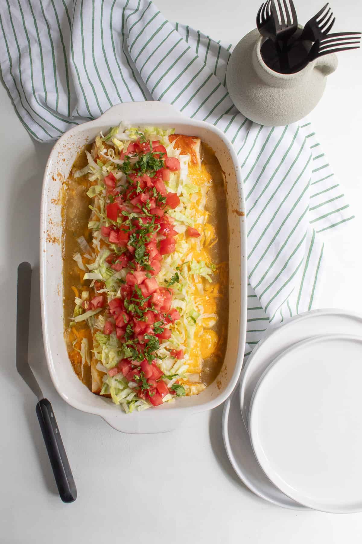 The finished casserole sits on a white surface next to a pile of plates, a spatula, and a container of forks.