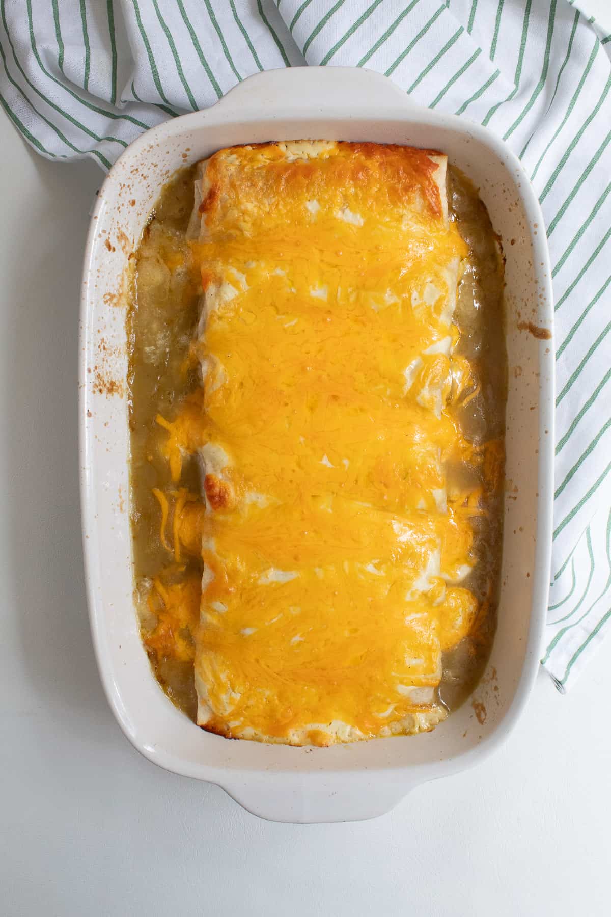 The baked casserole with melted cheese over the burritos sits on a white surface.