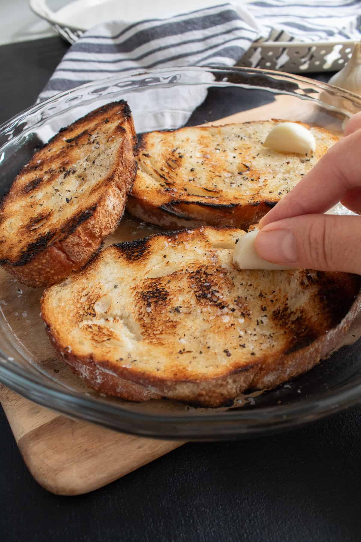 A clove of garlic is rubbed over the surface of a grilled piece of bread.