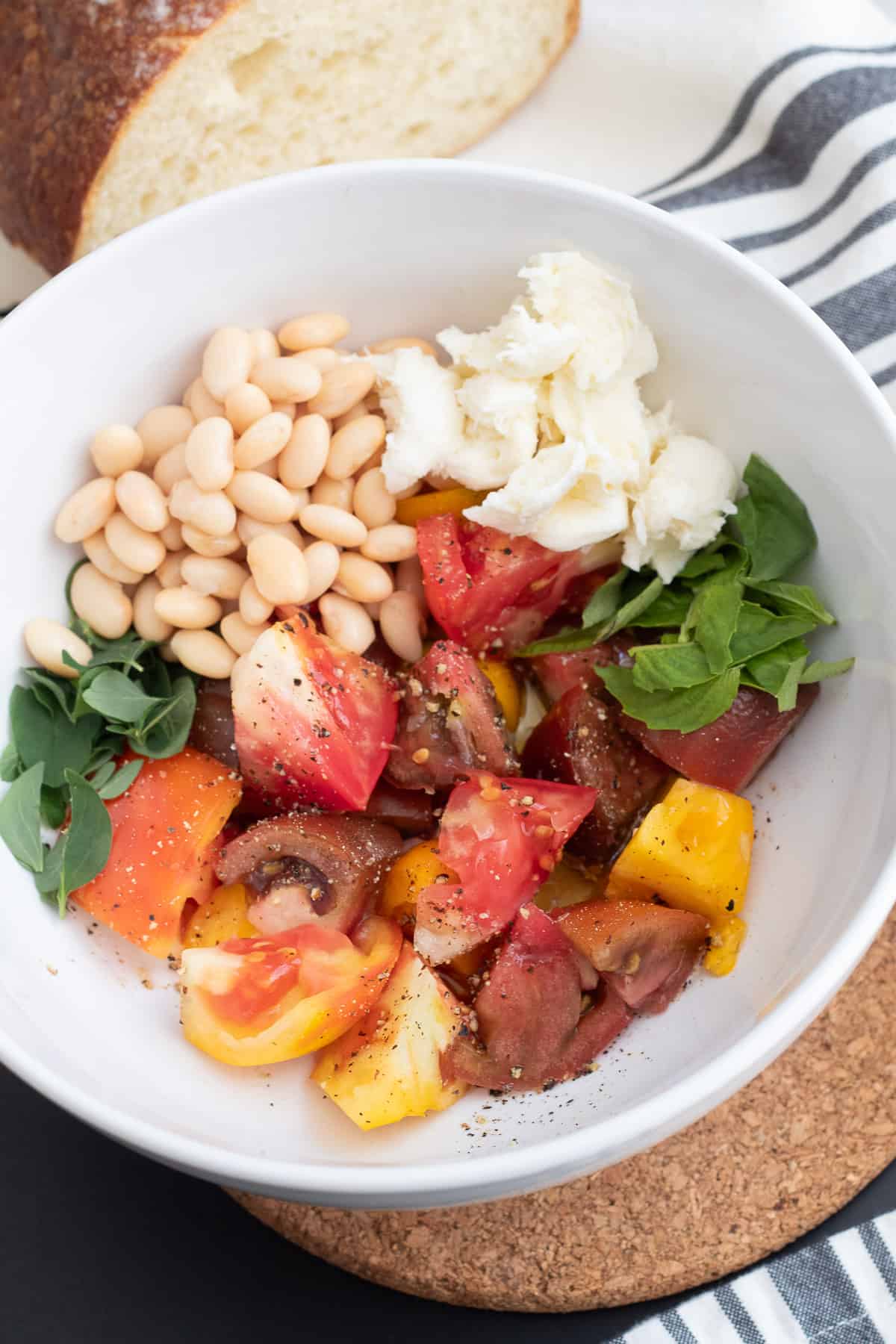 A white bowl contains piles of chopped tomato, herbs, cheese, and white beans and is dusted with salt and pepper.