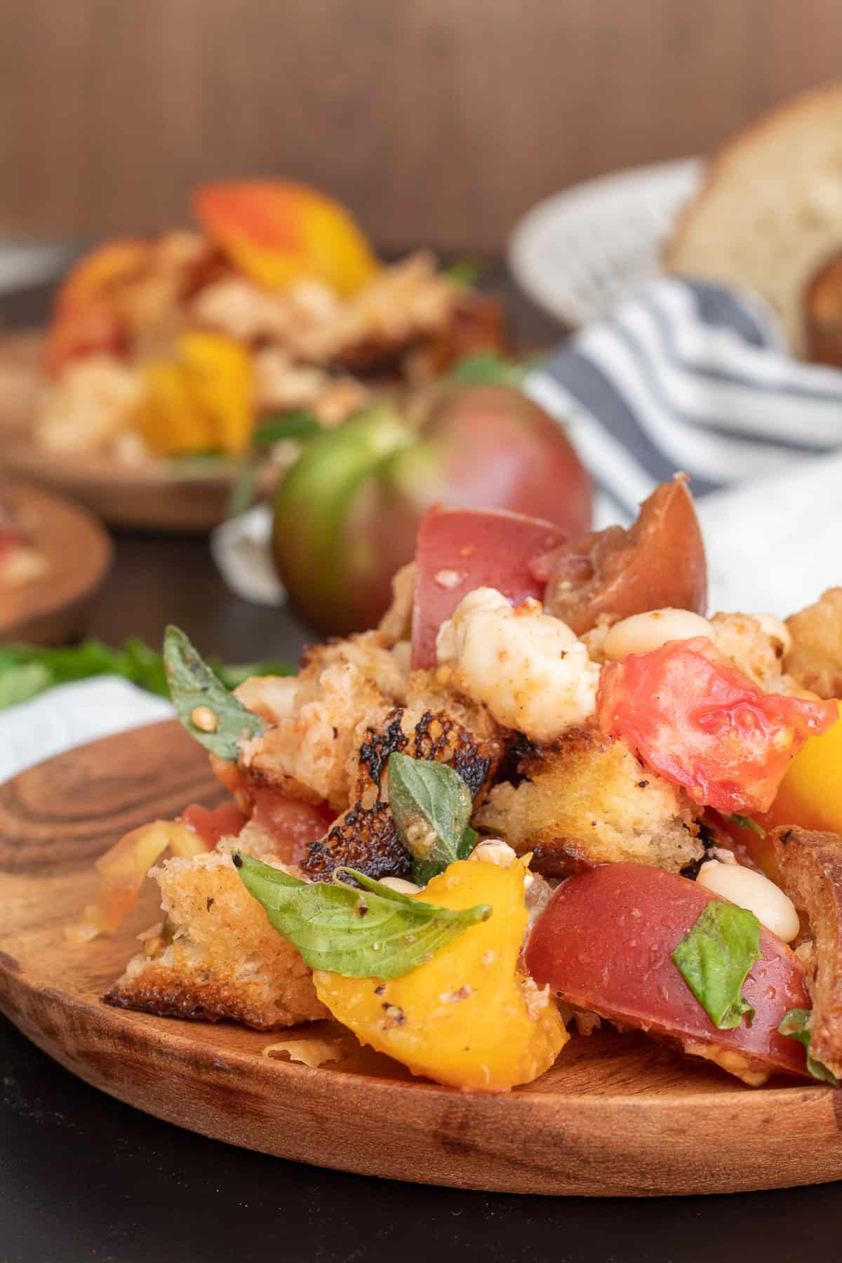 A wooden plate holds a detail image of a serving of the salad, with herbs, tomatoes, bread, beans, and cheese visible.