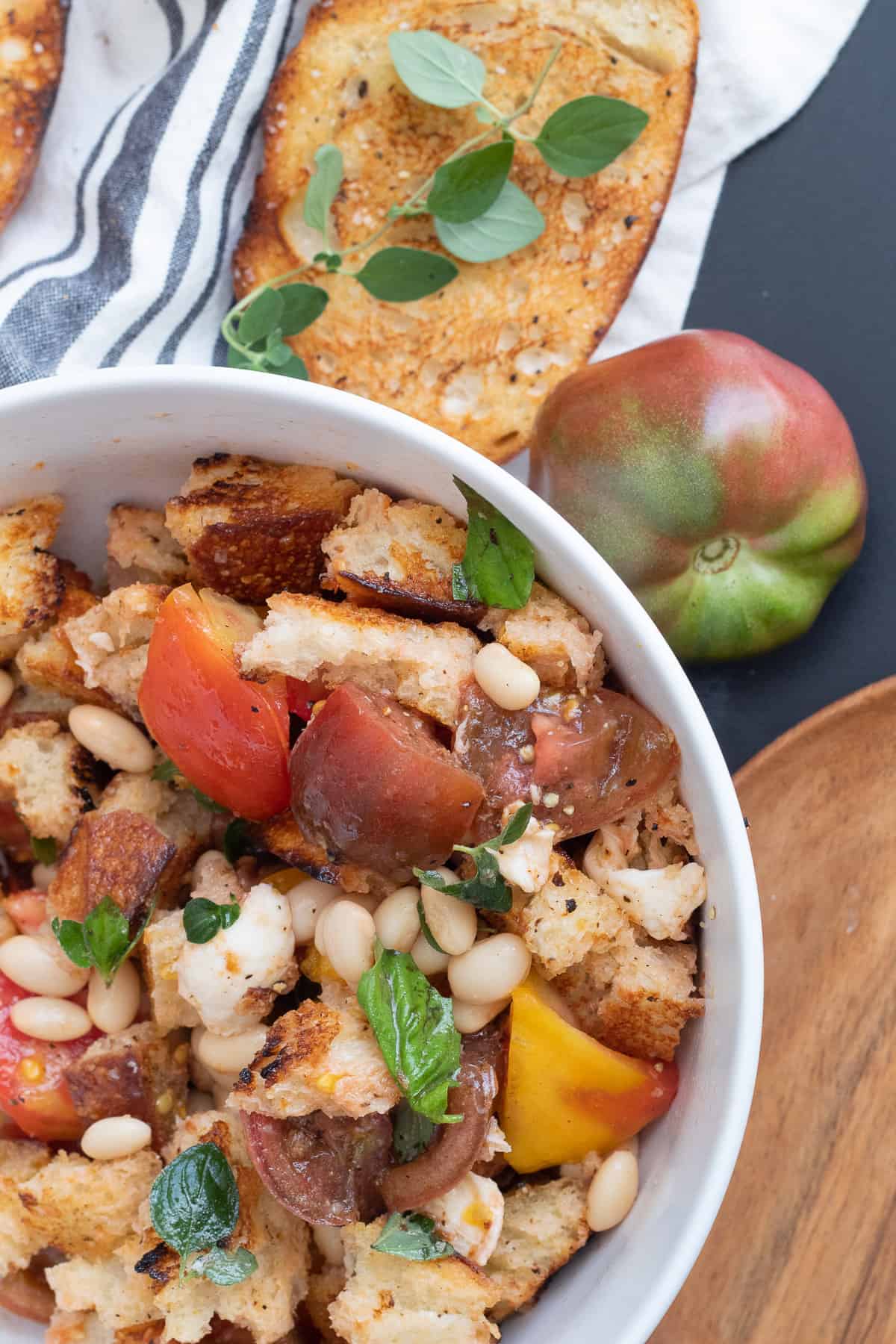 The mixed salad sits in a white bowl over a black surface next to a green and red tomato and oregano leaves.