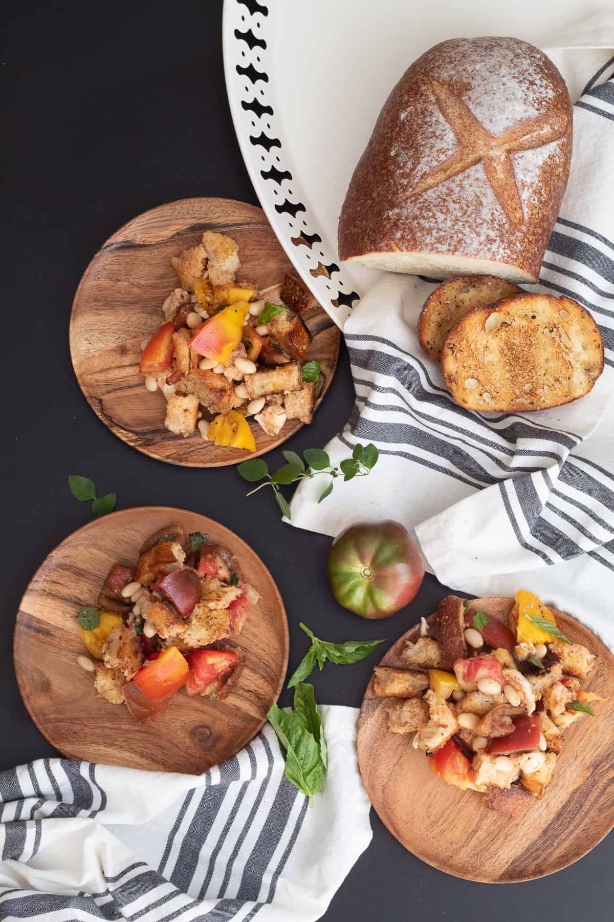 Three servings of the salad on wooden plates are arranged over a black surface with tomatoes, herbs, and grilled bread.