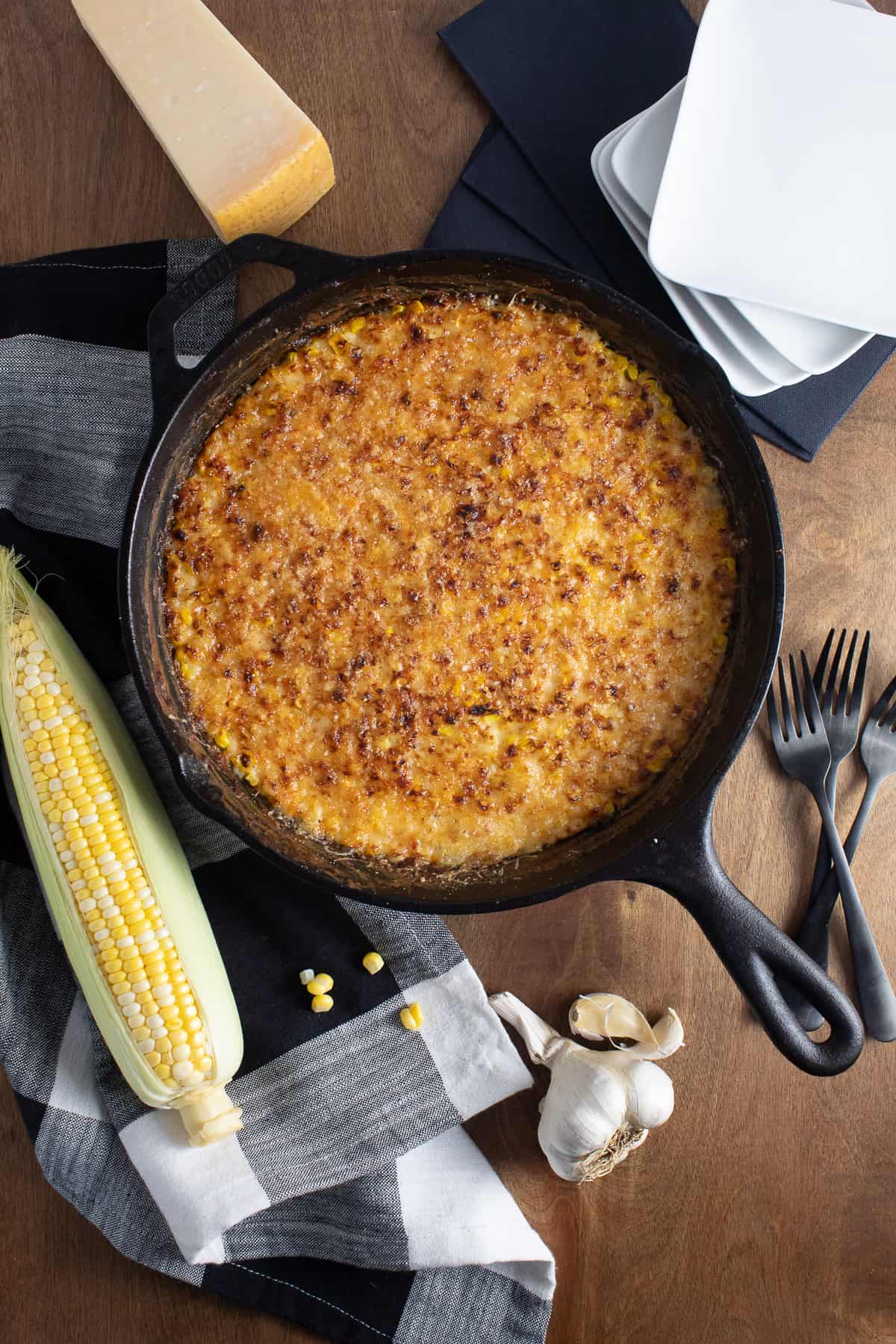 The bronzed gratin in a black cast iron skillet is at the center of the image, surrounded by plates, forks, an ear of corn, a Parmesan cheese wedge, and cloves of garlic.