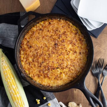 The bronzed gratin in a black cast iron skillet is at the center of the image, surrounded by plates, forks, an ear of corn, a Parmesan cheese wedge, and cloves of garlic.