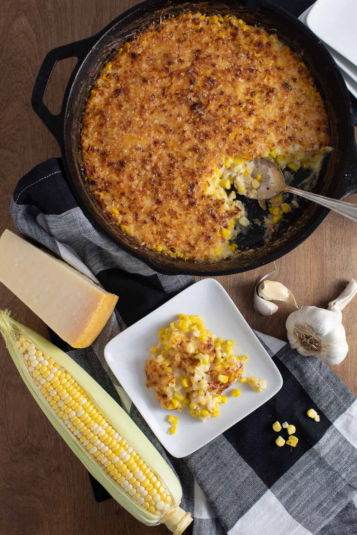 The finished casserole with its golden top is displayed next to a wedge of Parmesan cheese, an ear of corn, a serving of the gratin, and a bulb of garlic.