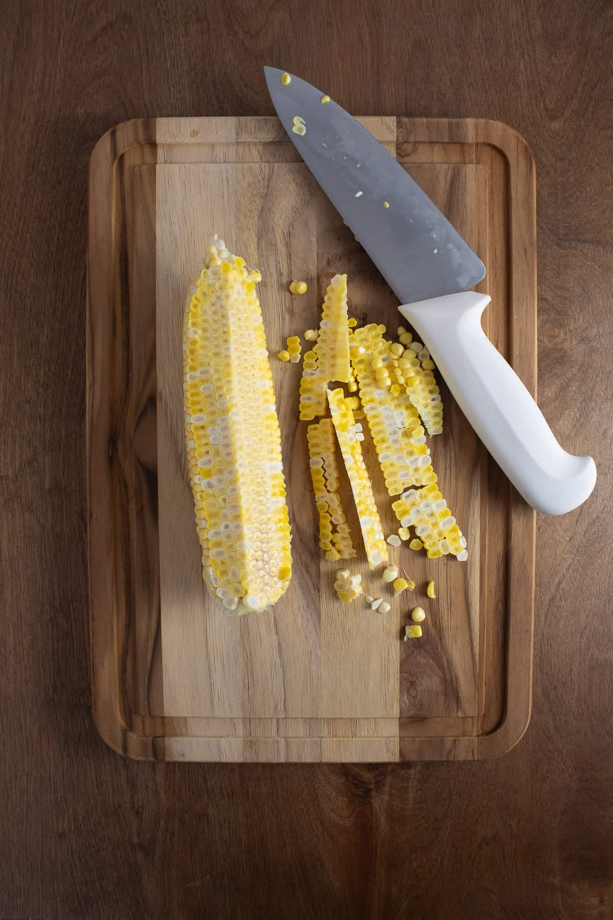 The kernels of corn being cut from the cob on a wooden cutting board with a white handled knife.