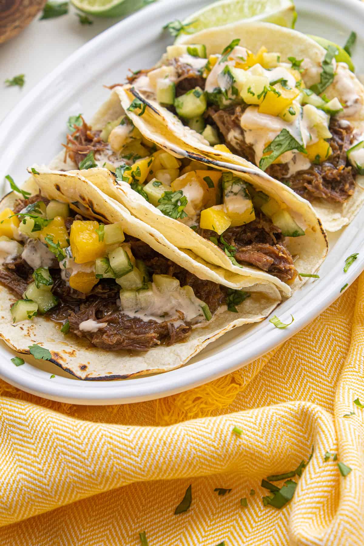 The plated tacos are displayed so that the shredded beef filling, sriracha mayo, and mango cucumber salad are visible.