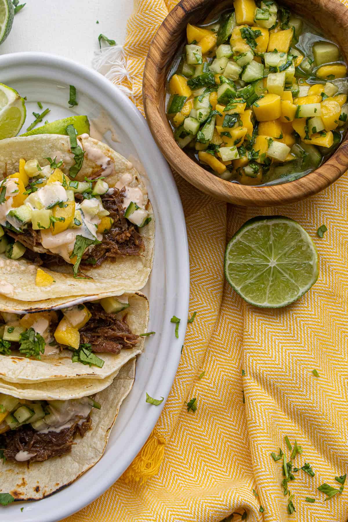 Plated tacos sit on a yellow napkin next to a halved lime and a bowl of salad for topping the tacos.