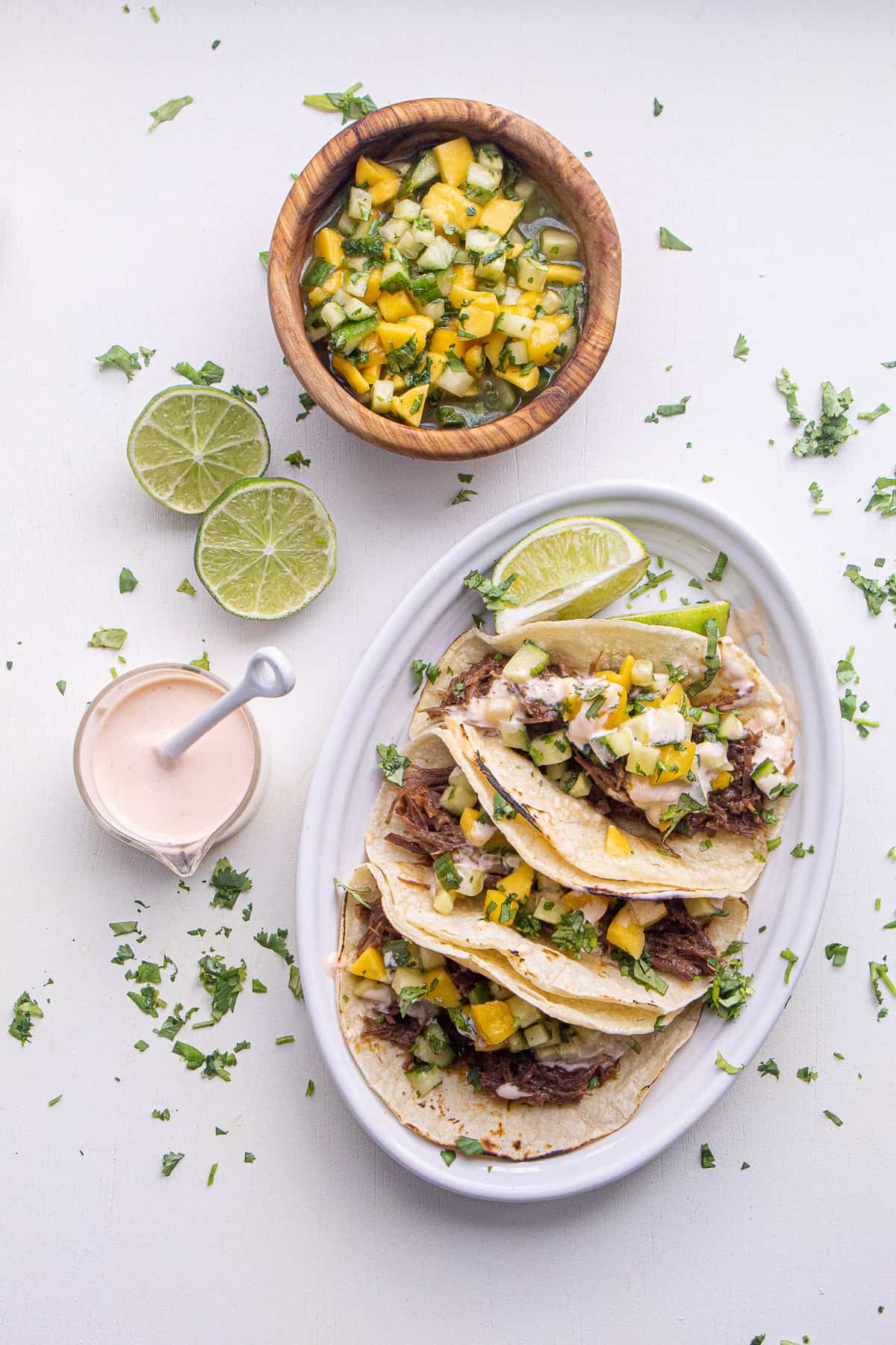 An oval plate of the tacos is sprinkled with cilantro and placed next to a small bowl of sriracha mayo, sliced limes, and mango cucumber salad.
