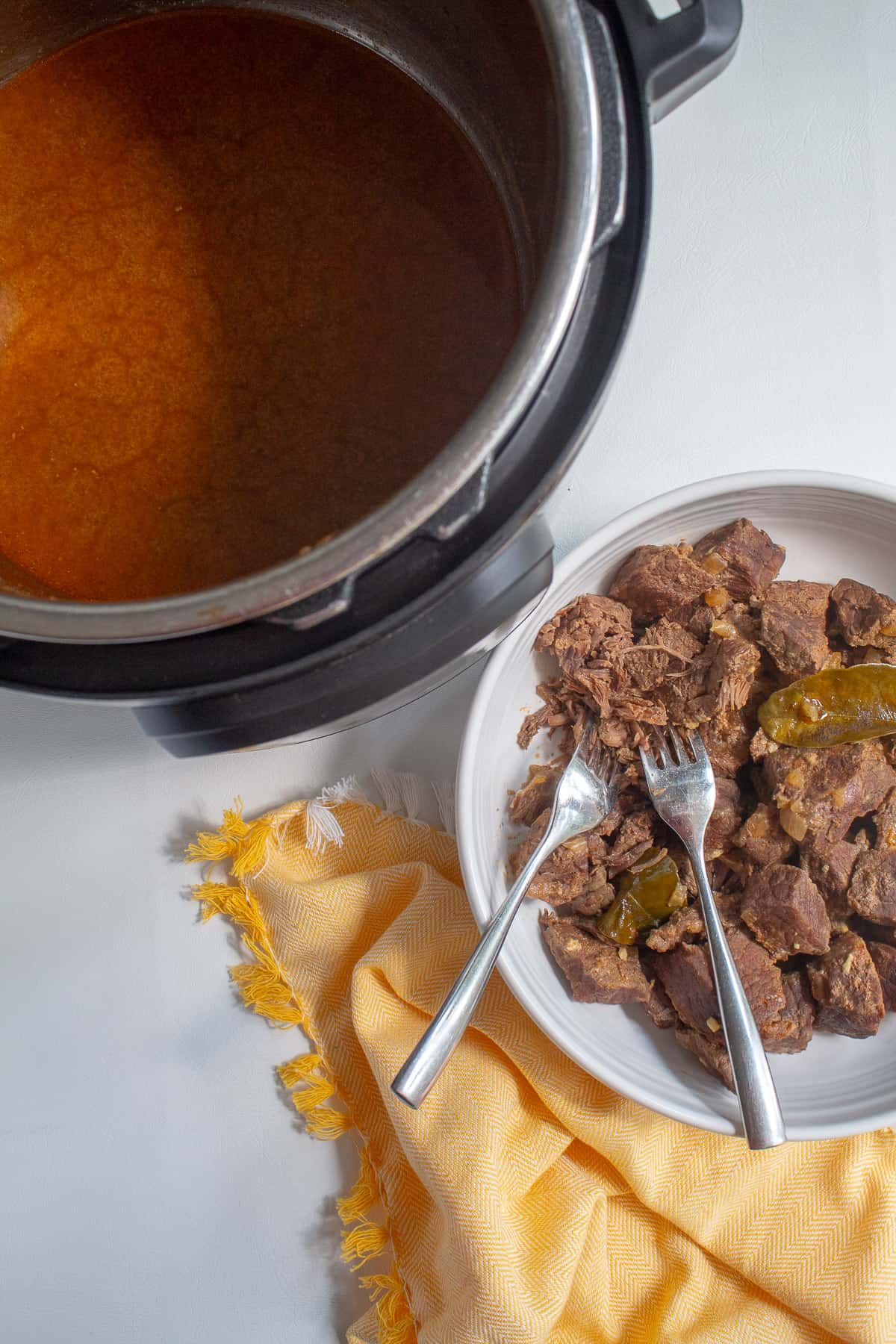 The cubes of cooked beef are in a white bowl with two forks for shredding and the Instant Pot filled with sauce sits nearby.