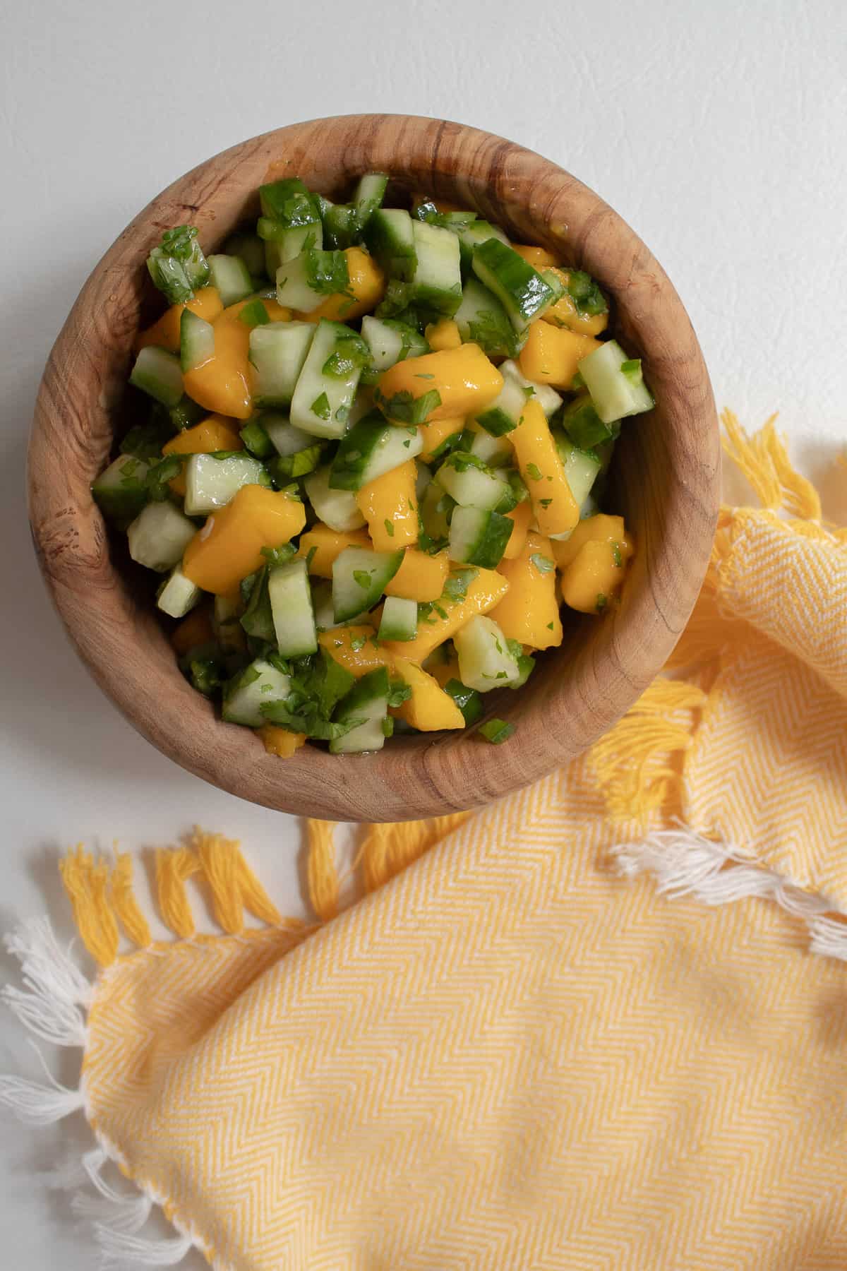 The finished mango cucumber salad is served in a small wooden bowl next to a pale yellow napkin.