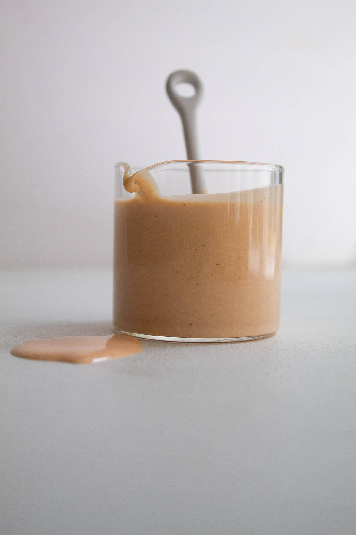 The sriracha mayo in a small spouted vessel sits on a white surface with the handle of a small spoon poking out the top.