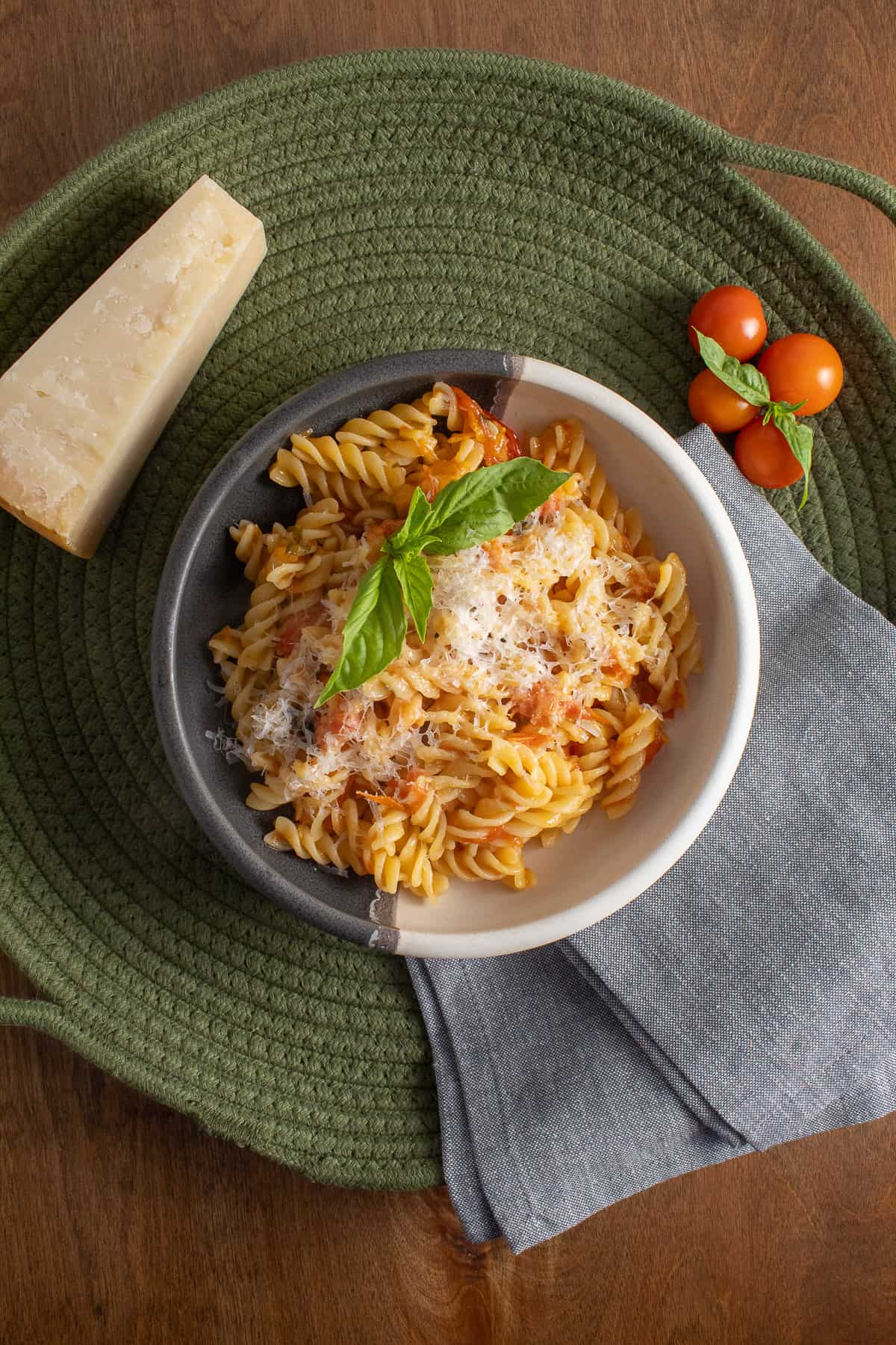 A gray and white bowl of fusilli pasta sits on a green woven tray near a wedge of Parmesan chees, cherry tomatoes, and basil.