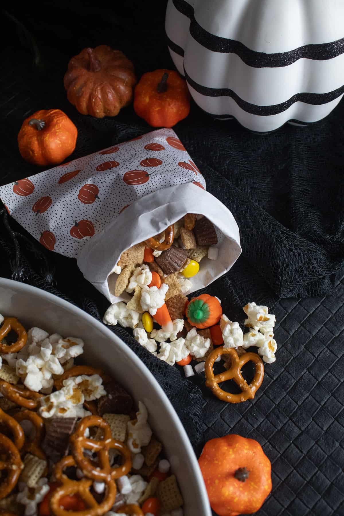 A paper bag with a pumpkin print is filled with the snack mix and spilling out on the black surface.